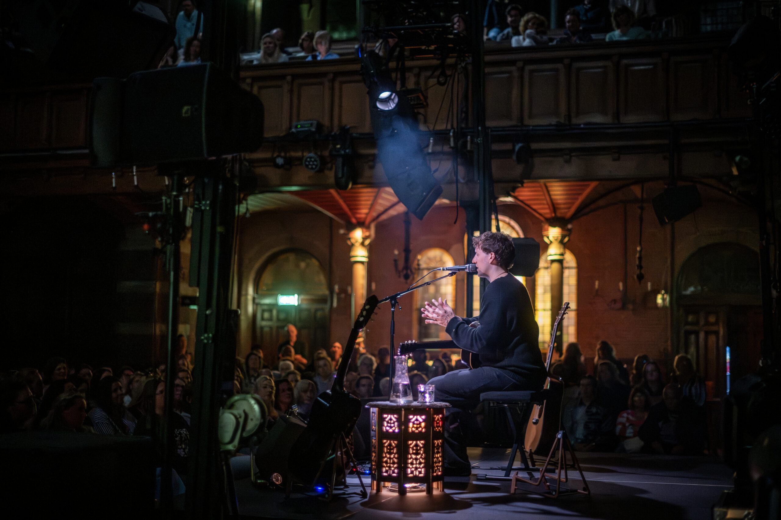 Zijaanzicht van het podium met publiek op de voorgrond en Eloi Youssef gezeten achter zijn tafeltje